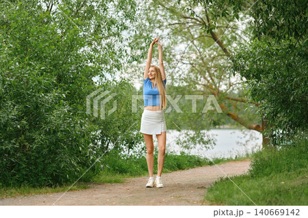 Young woman in sporty outfit stretches by path near water during daytime in green park Young woman in sporty outfit stretches by path near water during daytime in green park 140669142