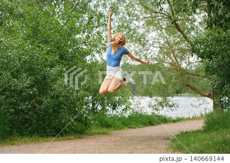 Sporty woman jumps joyfully along pathway near water surrounded by green trees on sunny day Sporty woman jumps joyfully along pathway near water surrounded by green trees on sunny day 140669144