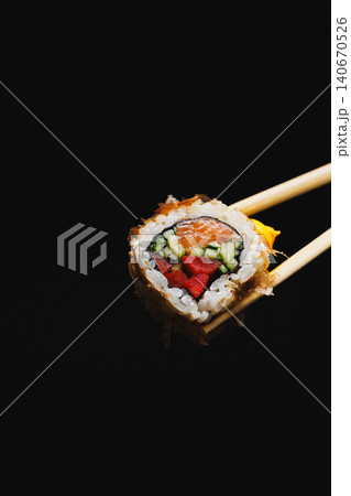 Fresh sushi roll with salmon, cucumber, and strawberry held by chopsticks against a black background 140670526