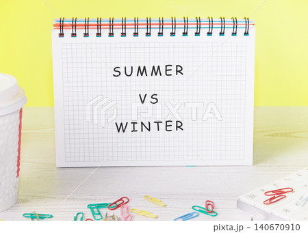 A notebook with the words summer vs winter is placed on a table. Colorful paper clips are scattered on the surface. The background is bright yellow 140670910