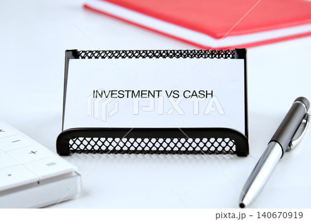 A card shows the words investment vs cash in a financial workspace. A calculator and pen are nearby, along with a red notebook. The setting suggests planning or analysis A card shows the words investment vs cash in a financial workspace. A calculator and pen are nearby, along with a red notebook. The setting suggests planning or analysis 140670919