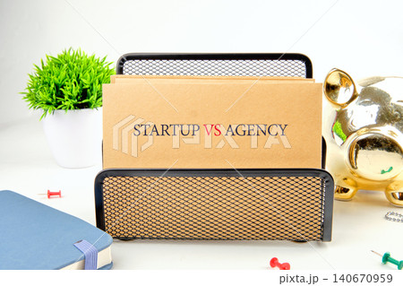 Two cards show the words startup and agency on a desk with a plant, a piggy bank, and a notebook. This scene takes place in a bright office environment 140670959