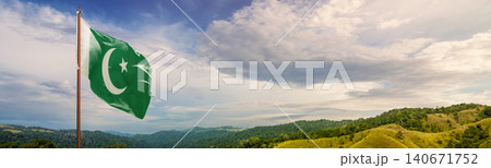The national flag of Pakistan fluttering in the wind on a flagpole with a panoramic background of green hills and a cloudy blue sky 140671752