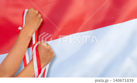 Two raised fists hold red and white ribbons against a large Indonesian flag, symbolizing independence, strength, and national pride. Hari Kemerdekaan Dirgahayu Indonesia Two raised fists hold red and white ribbons against a large Indonesian flag, symbolizing independence, strength, and national pride. Hari Kemerdekaan Dirgahayu Indonesia 140671839