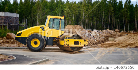 Yellow road roller compactor parked on a construction site with piles of earth and trees in the background, showcasing heavy machinery in action 140672550