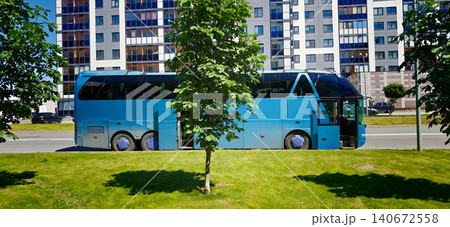 Blue tour bus parked beside a green lawn with trees in front of a modern residential building featuring large windows and a clear blue sky Blue tour bus parked beside a green lawn with trees in front of a modern residential building featuring large windows and a clear blue sky 140672558