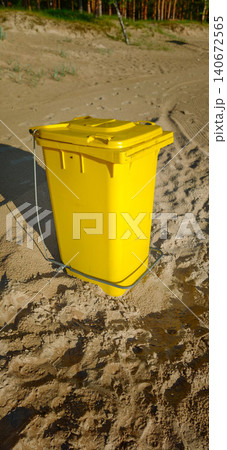 Yellow waste bin positioned on sandy beach with natural vegetation and trees visible in the background under bright sunlight 140672565