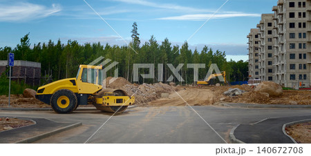 Yellow road roller and excavator on construction site with unfinished building and forest in the background under a clear blue sky Yellow road roller and excavator on construction site with unfinished building and forest in the background under a clear blue sky 140672708