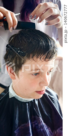Young boy receiving a haircut at a salon, stylist using a comb to style hair while wearing a black cape, with soft lighting illuminating the scene 140672777