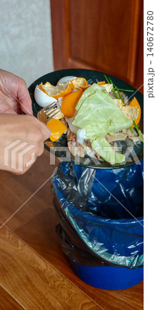 Hand holding a plate of food scraps over a blue trash bin, with various vegetable peels and leftovers ready for disposal in a kitchen setting Hand holding a plate of food scraps over a blue trash bin, with various vegetable peels and leftovers ready for disposal in a kitchen setting 140672780
