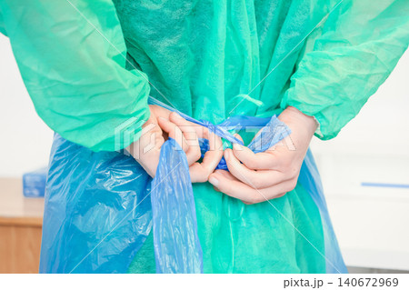 Medical professional hands tying blue apron strings behind back while wearing protective green gown, conveying preparation and safety protocols in a busy sterile clinical health environment 140672969