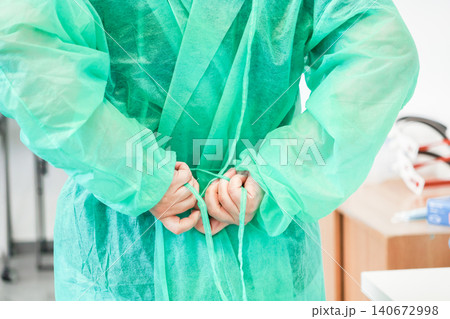 A medical professional ties green protective gown strings behind their back at a hospital clinic, representing essential hygiene preparation for a sterile healthcare safety environment 140672998