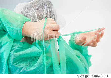 A medical professional ties a green surgical gown in a sterile hospital setting, preparing for a procedure while ensuring proper protective equipment and hygiene safety standards A medical professional ties a green surgical gown in a sterile hospital setting, preparing for a procedure while ensuring proper protective equipment and hygiene safety standards 140673001
