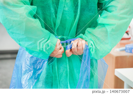 A dedicated medical professional ties a green disposable surgical gown behind their back in a sterile clinic to emphasize strict safety protocols and infection control in hospitals A dedicated medical professional ties a green disposable surgical gown behind their back in a sterile clinic to emphasize strict safety protocols and infection control in hospitals 140673007