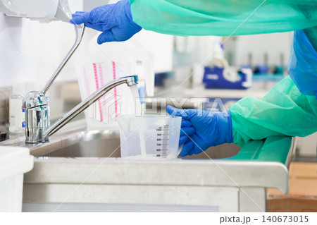 Gloved hands carefully filling a clear plastic beaker with water in a clinical lab setting to emphasize professional sanitation and rigorous safety standards for medical research Gloved hands carefully filling a clear plastic beaker with water in a clinical lab setting to emphasize professional sanitation and rigorous safety standards for medical research 140673015