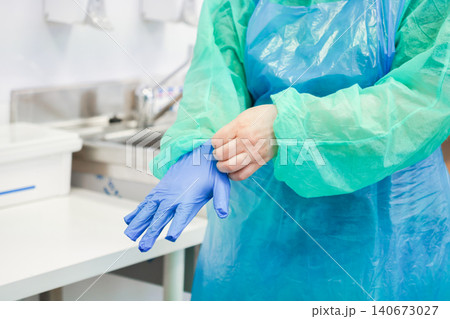 A medical professional puts on a blue nitrile glove while wearing a protective gown in a clinical hospital setting to highlight hygiene standards and safety protocols for care 140673027