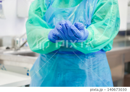 A medical professional puts on blue nitrile gloves in a clinic while wearing a protective gown to prepare for a clinical procedure ensuring safety and hygiene in a healthcare setting A medical professional puts on blue nitrile gloves in a clinic while wearing a protective gown to prepare for a clinical procedure ensuring safety and hygiene in a healthcare setting 140673030