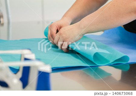 Medical professional hands folding sterile blue surgical paper on a metallic table, representing hygiene, clinical preparation, equipment maintenance, and safety in hospitals Medical professional hands folding sterile blue surgical paper on a metallic table, representing hygiene, clinical preparation, equipment maintenance, and safety in hospitals 140673068