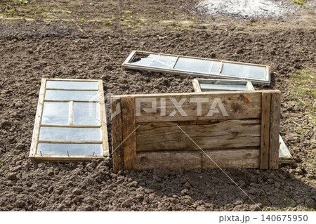 Old wooden window frames and panels resting on dark plowed soil in a garden, creating a rustic textured scene that highlights abandoned materials and preparation for spring planting Old wooden window frames and panels resting on dark plowed soil in a garden, creating a rustic textured scene that highlights abandoned materials and preparation for spring planting 140675950