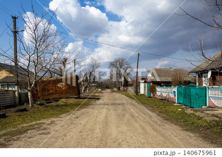 Unpaved dirt road leading through a quiet rural village with traditional small houses and bare trees under a bright blue sky, representing simple countryside life and remote areas Unpaved dirt road leading through a quiet rural village with traditional small houses and bare trees under a bright blue sky, representing simple countryside life and remote areas 140675961