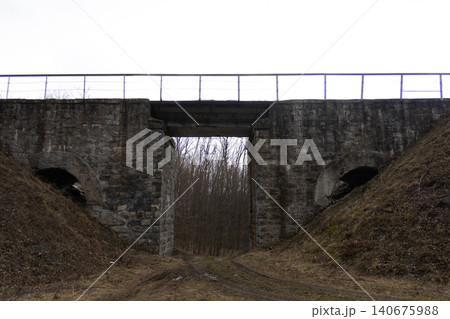 An old stone railway bridge spans a forest path with arched brick construction that creates a rustic passage through nature, evoking a quiet sense of historical transportation An old stone railway bridge spans a forest path with arched brick construction that creates a rustic passage through nature, evoking a quiet sense of historical transportation 140675988