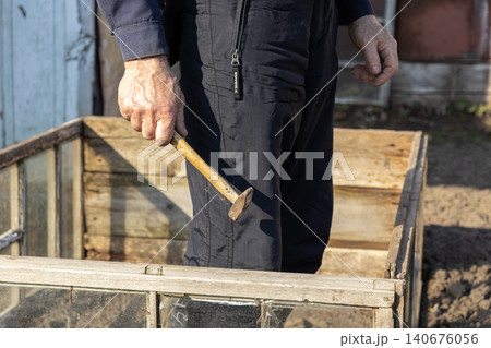 A senior man holds a vintage hammer over a wooden garden frame, representing manual labor, small home improvement projects, and active outdoor property maintenance during the dusk A senior man holds a vintage hammer over a wooden garden frame, representing manual labor, small home improvement projects, and active outdoor property maintenance during the dusk 140676056
