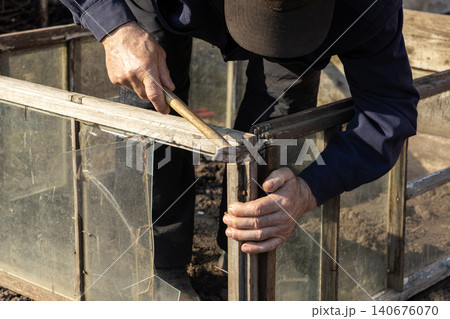 A skilled person uses a metal hammer to repair an old wooden garden greenhouse frame, reflecting the manual labor and dedication needed for seasonal outdoor home improvement tasks A skilled person uses a metal hammer to repair an old wooden garden greenhouse frame, reflecting the manual labor and dedication needed for seasonal outdoor home improvement tasks 140676070