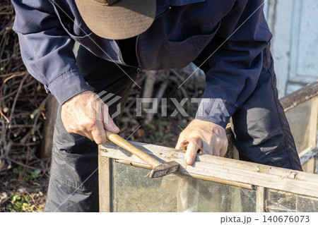 Person hammering a nail into an old wooden window frame, focusing on a home renovation task outdoors to showcase manual labor, repair skills, and craftsmanship in a rustic setting Person hammering a nail into an old wooden window frame, focusing on a home renovation task outdoors to showcase manual labor, repair skills, and craftsmanship in a rustic setting 140676073