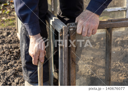 A person carefully assembles a wooden garden cold frame with glass panels over soil to protect plants, creating an organized outdoor space for spring planting and home gardening A person carefully assembles a wooden garden cold frame with glass panels over soil to protect plants, creating an organized outdoor space for spring planting and home gardening 140676077