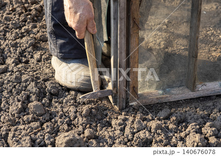 An older person uses a metal hammer to repair a vintage wooden greenhouse frame in the soil, highlighting traditional DIY craftsmanship during a bright sunny outdoor project An older person uses a metal hammer to repair a vintage wooden greenhouse frame in the soil, highlighting traditional DIY craftsmanship during a bright sunny outdoor project 140676078