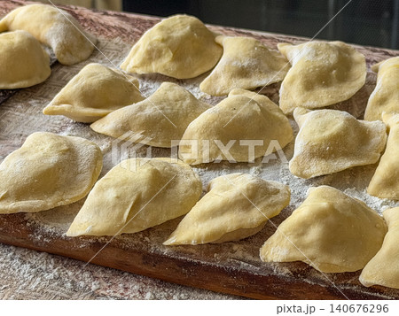 Raw handmade dumplings arranged on a floured wooden cutting board, capturing the authentic process of preparing traditional dough filled parcels for a delicious homemade meal project Raw handmade dumplings arranged on a floured wooden cutting board, capturing the authentic process of preparing traditional dough filled parcels for a delicious homemade meal project 140676296