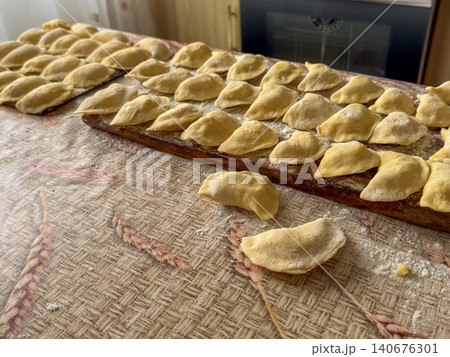 Fresh handmade dumplings arranged on a wooden board across a kitchen table, capturing the traditional process of preparing authentic homemade dough food for a rustic meal experience Fresh handmade dumplings arranged on a wooden board across a kitchen table, capturing the traditional process of preparing authentic homemade dough food for a rustic meal experience 140676301