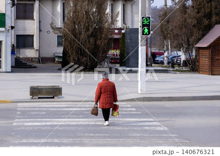 A woman in a warm orange jacket walks across a town square pedestrian crossing while a green signal counts down, illustrating safe daily urban transit in a cold morning setting 140676321