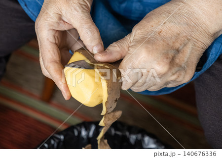 A senior person peels a raw potato with a small steel knife, representing traditional home cooking preparation and domestic life in a serene atmosphere of culinary skill and care A senior person peels a raw potato with a small steel knife, representing traditional home cooking preparation and domestic life in a serene atmosphere of culinary skill and care 140676336