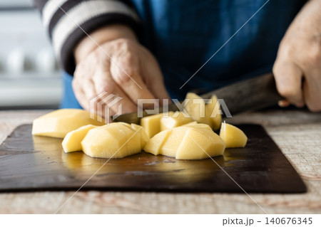 A senior person carefully cuts a raw peeled potato on a dark kitchen board, illustrating culinary preparation and healthy homemade cooking techniques for delicious nutritious meals 140676345