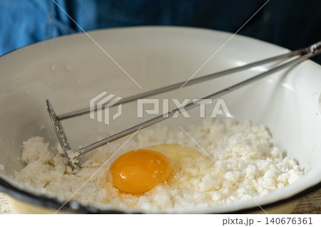 Fresh cottage cheese with a raw egg yolk in a white bowl being mashed with a metal tool for a homemade dairy baking preparation in a bright kitchen setting for healthy cooking recipes 140676361