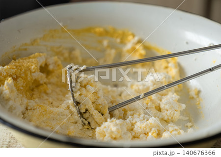 A manual potato masher crushing cottage cheese with egg yolk in a rustic white bowl, capturing a home cooking preparation process for delicious healthy protein snacks and light meals A manual potato masher crushing cottage cheese with egg yolk in a rustic white bowl, capturing a home cooking preparation process for delicious healthy protein snacks and light meals 140676366