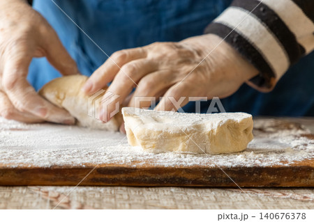 An elderly person shapes soft raw dough on a wooden board covered in white flour, representing traditional artisanal baking techniques for a cozy homemade meal in the rustic kitchen An elderly person shapes soft raw dough on a wooden board covered in white flour, representing traditional artisanal baking techniques for a cozy homemade meal in the rustic kitchen 140676378
