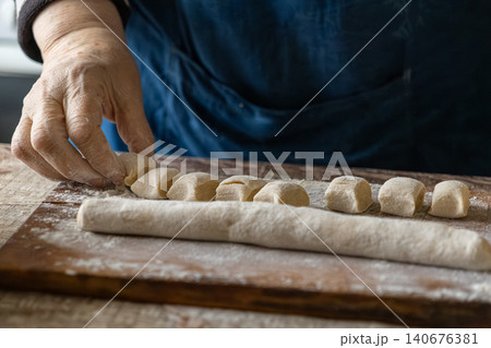 A pair of senior hands cuts pieces of raw dough on a wooden board to create handmade gnocchi, emphasizing authentic culinary traditions and a nurturing home cooking kitchen atmosphere A pair of senior hands cuts pieces of raw dough on a wooden board to create handmade gnocchi, emphasizing authentic culinary traditions and a nurturing home cooking kitchen atmosphere 140676381