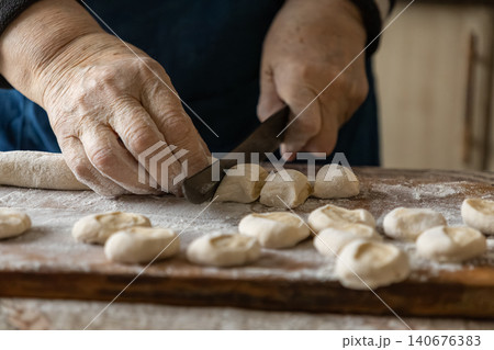 Elderly person hand cutting fresh dough into small pieces with a sharp knife on wooden board, showcasing traditional dumpling preparation for authentic homemade culinary cooking craft Elderly person hand cutting fresh dough into small pieces with a sharp knife on wooden board, showcasing traditional dumpling preparation for authentic homemade culinary cooking craft 140676383