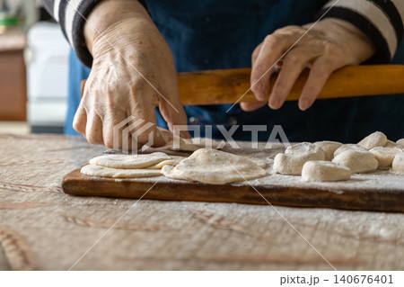 An older person rolls fresh dough circles on a wooden board to prepare traditional dumplings, conveying authentic home cooking skills and a warm cultural atmosphere in the kitchen An older person rolls fresh dough circles on a wooden board to prepare traditional dumplings, conveying authentic home cooking skills and a warm cultural atmosphere in the kitchen 140676401