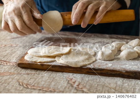 Senior person rolls fresh dough circles with a wooden pin on a floured board to prepare homemade dumplings, capturing the traditional craft of cooking healthy and authentic food Senior person rolls fresh dough circles with a wooden pin on a floured board to prepare homemade dumplings, capturing the traditional craft of cooking healthy and authentic food 140676402