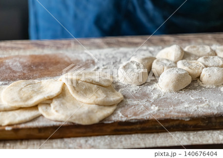 Fresh dough balls and rolled flat discs sitting on a floured wooden board, representing traditional dumpling preparation in a rustic kitchen setting focused on homemade meal cooking Fresh dough balls and rolled flat discs sitting on a floured wooden board, representing traditional dumpling preparation in a rustic kitchen setting focused on homemade meal cooking 140676404