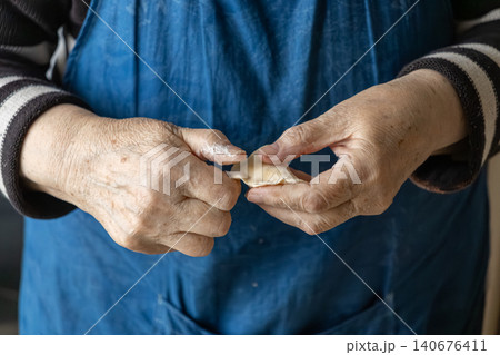 A senior person carefully shaping a handmade dumpling with floured fingers in a blue apron, capturing the traditional culinary craft and authentic home cooking process in a kitchen A senior person carefully shaping a handmade dumpling with floured fingers in a blue apron, capturing the traditional culinary craft and authentic home cooking process in a kitchen 140676411