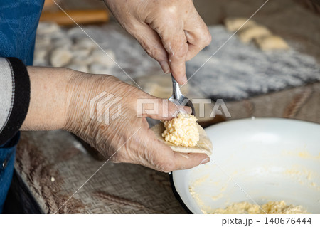 A senior person preparing homemade cottage cheese dumplings by placing creamy filling onto circular dough circles with a small metal spoon inside a cozy rustic kitchen home setting A senior person preparing homemade cottage cheese dumplings by placing creamy filling onto circular dough circles with a small metal spoon inside a cozy rustic kitchen home setting 140676444