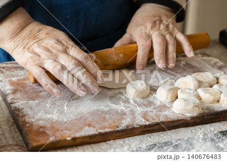 An elderly person uses a wooden rolling pin to flatten dough for fresh dumplings on a floured kitchen board, representing authentic culinary tradition and homemade comfort food craft 140676483