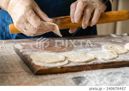 A senior person uses a wooden rolling pin to shape fresh dumpling dough on a dusted board, capturing a traditional culinary process and the art of handmade food in a warm kitchen A senior person uses a wooden rolling pin to shape fresh dumpling dough on a dusted board, capturing a traditional culinary process and the art of handmade food in a warm kitchen 140676487