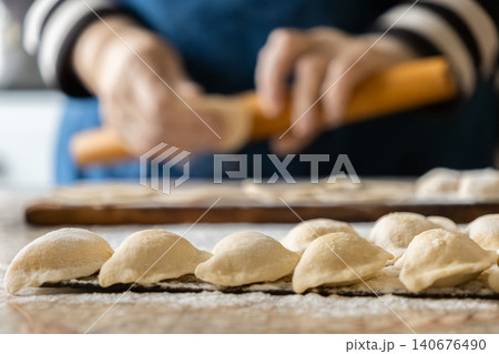 A chef prepares traditional dumplings by rolling dough on a floured wooden surface, capturing the authentic essence of handmade craft in a warm and cozy home kitchen environment A chef prepares traditional dumplings by rolling dough on a floured wooden surface, capturing the authentic essence of handmade craft in a warm and cozy home kitchen environment 140676490