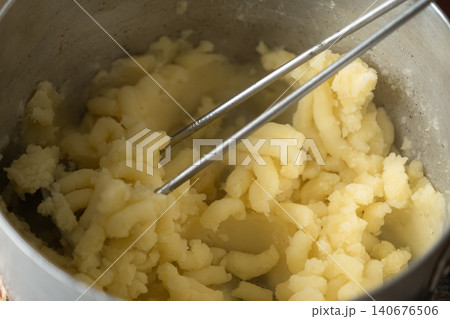 Boiled potato being mashed inside a metallic pot with a steel tool to prepare a filling for handmade savory dumplings, showcasing the rustic cooking process in a home kitchen Boiled potato being mashed inside a metallic pot with a steel tool to prepare a filling for handmade savory dumplings, showcasing the rustic cooking process in a home kitchen 140676506