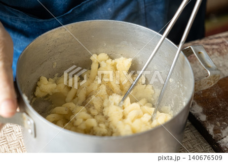 A chef mashes boiled potatoes in a metal pot with a wire masher to prepare dumpling filling, capturing the rustic culinary process of making traditional handmade dough for meals 140676509